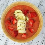 A smoothie bowl served in a yellow bowl with sliced bananas, chopped strawberries and chia seed
