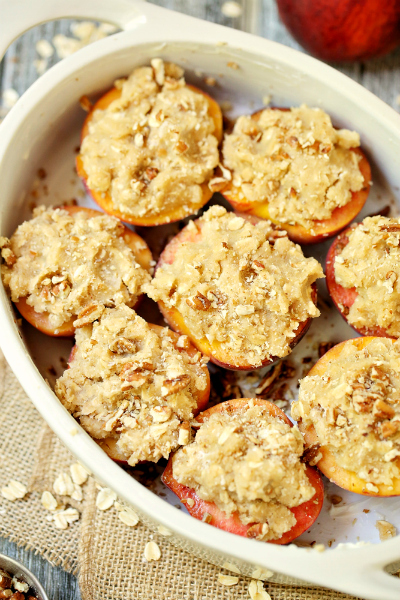 Halves of peaches sitting, topped with oat streusel topping, in a baking white baking dish