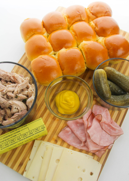 Ingredients for cuban sliders on a cutting board - sheet buns, pickles, mustard, butter, ham, sliced slice cheese and stick of butter
