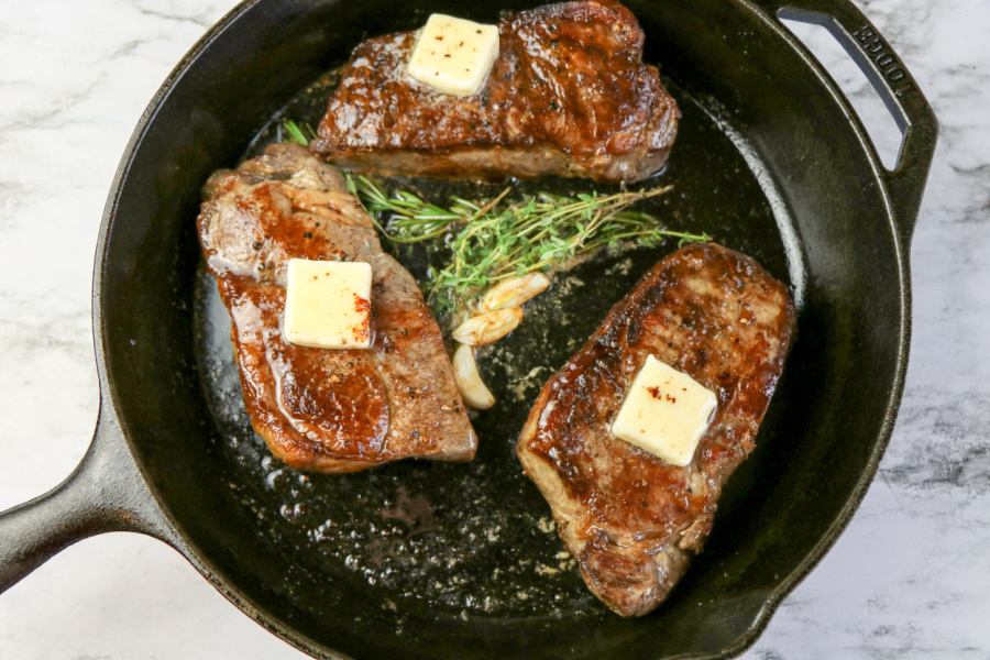3 steaks cooking in a cast iron skillet with herbs