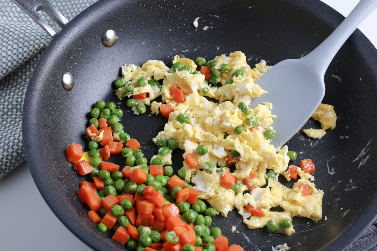 scrambled eggs and peas and carrots being mixed together in pan